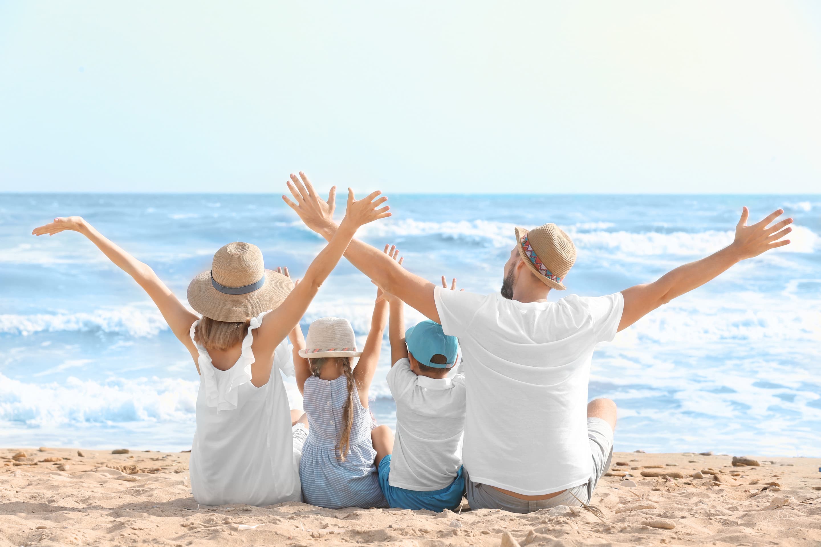 Four people, two adults and two children, sit on the beach facing the ocean with their arms raised, wearing hats and casual summer clothing.