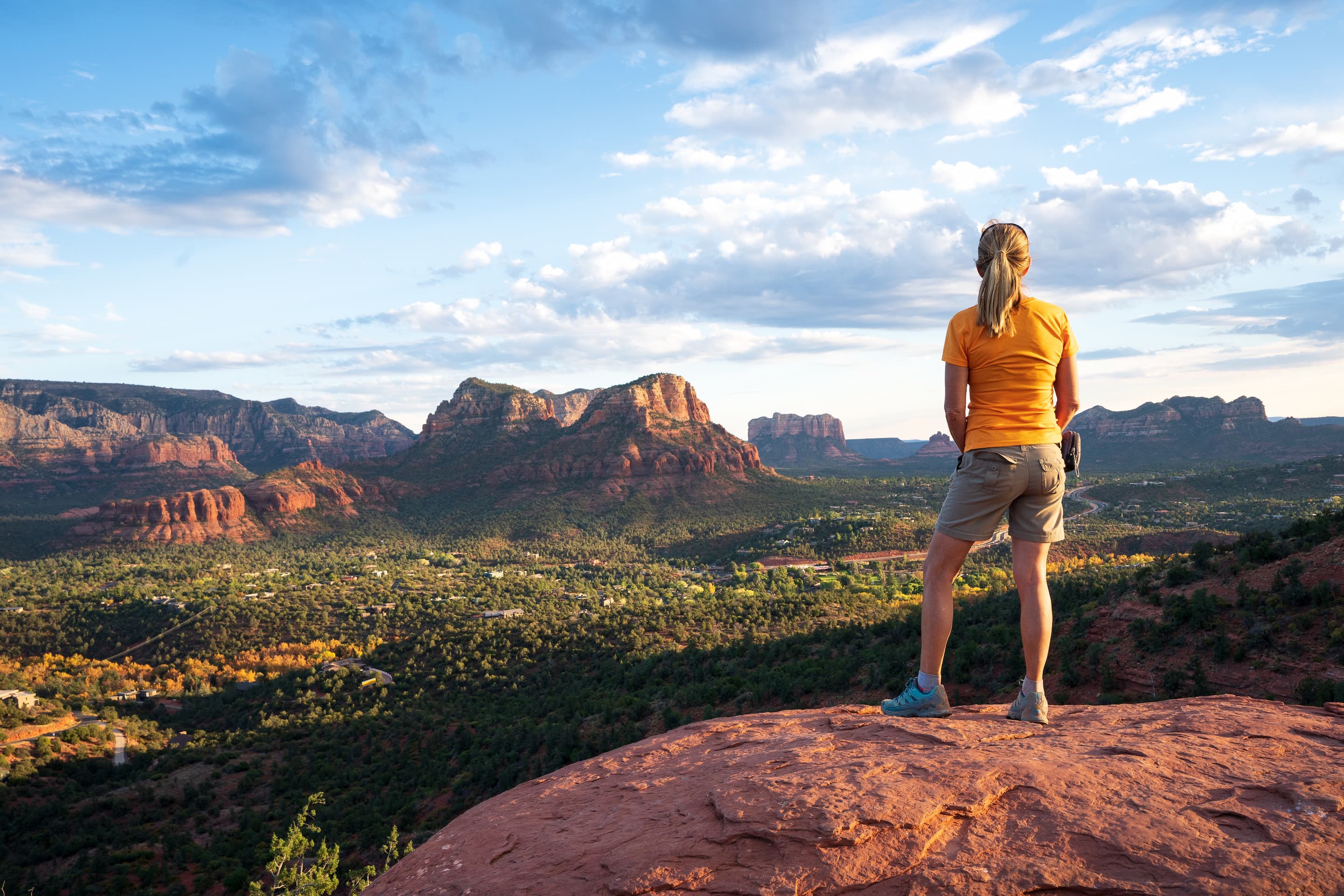 A person in an orange shirt stands on a rocky ledge, overlooking a vast landscape of red rock formations and green valleys under a partly cloudy sky.