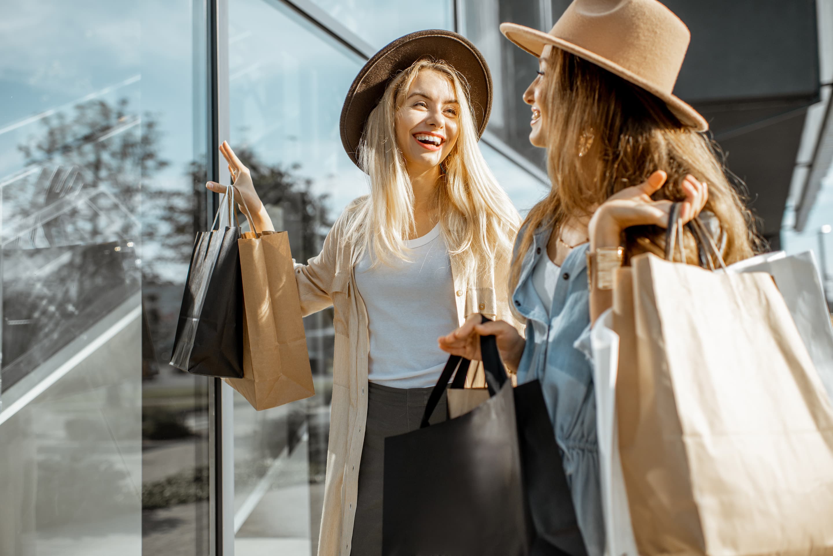 Two women wearing hats and holding shopping bags stand outside a store, smiling and talking to each other.