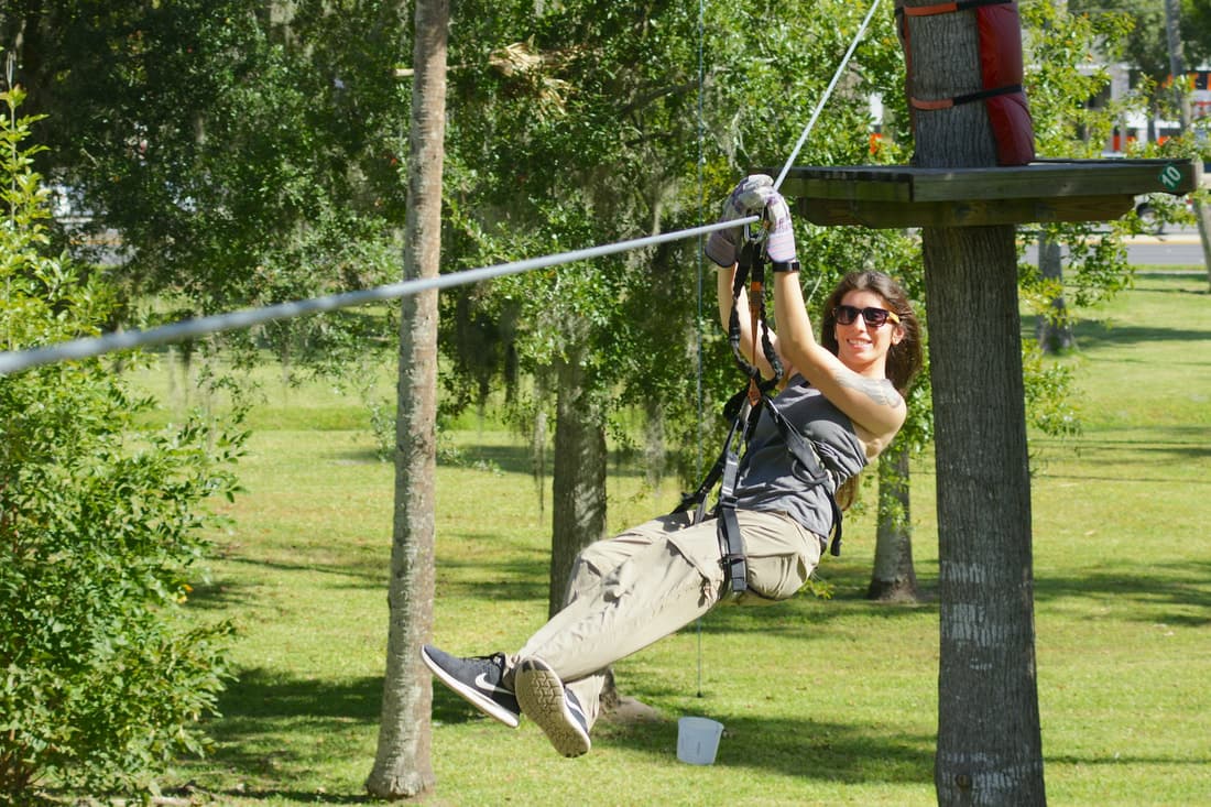A person wearing sunglasses and a harness rides a zip line outdoors, surrounded by trees and grass on a sunny day.
